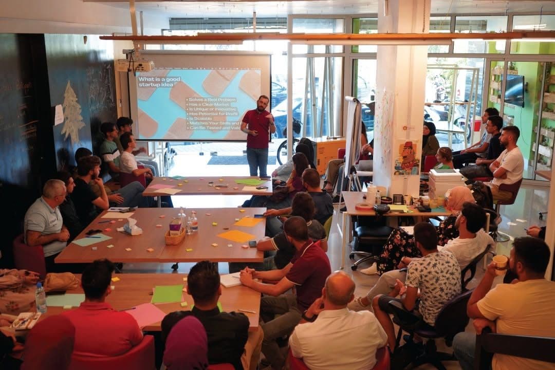 A trainer stands at a projected slide while participants work around tables