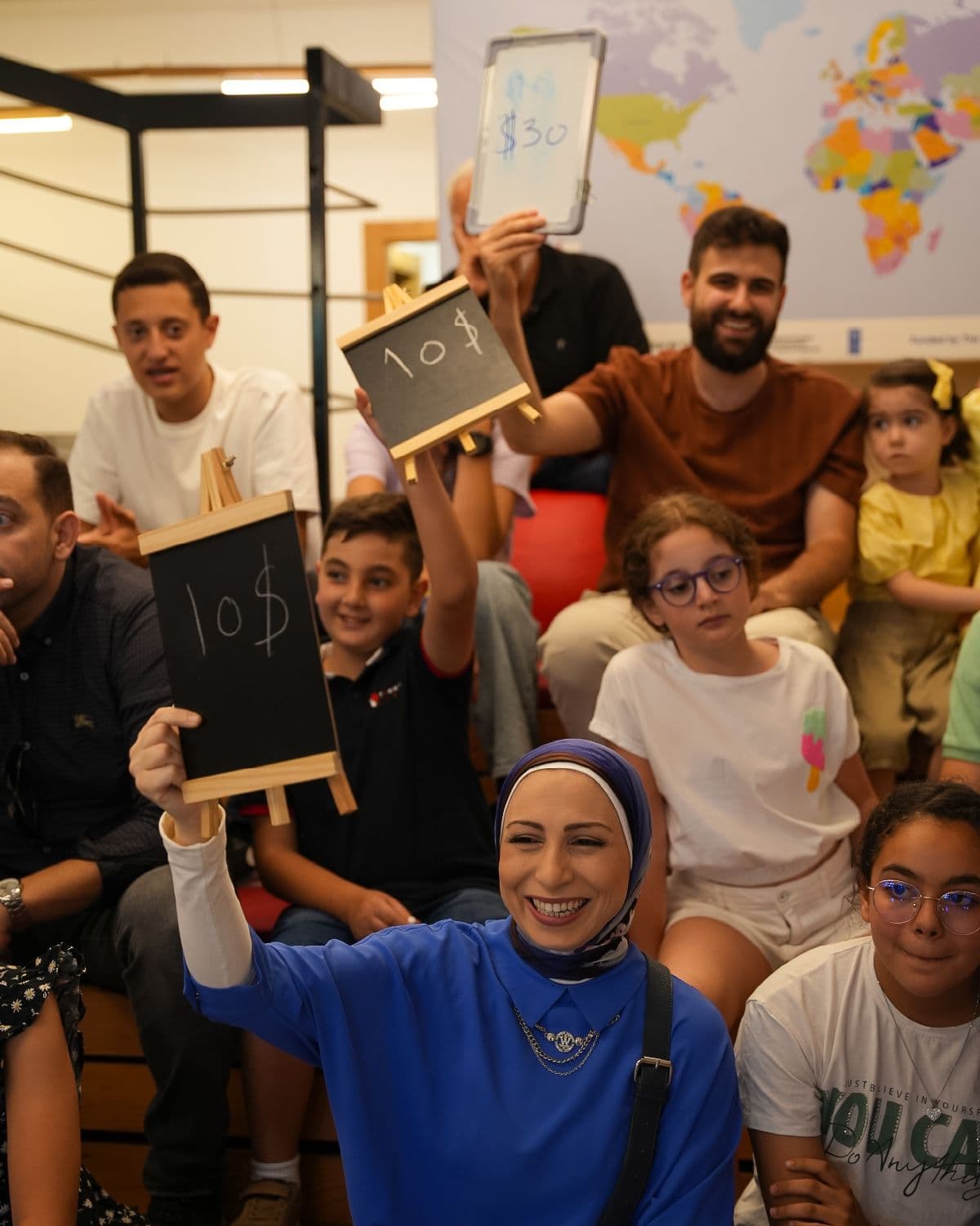 Parents and children in the audience hold up small chalkboards with numbers written on them during a Youngpreneurs Academy session