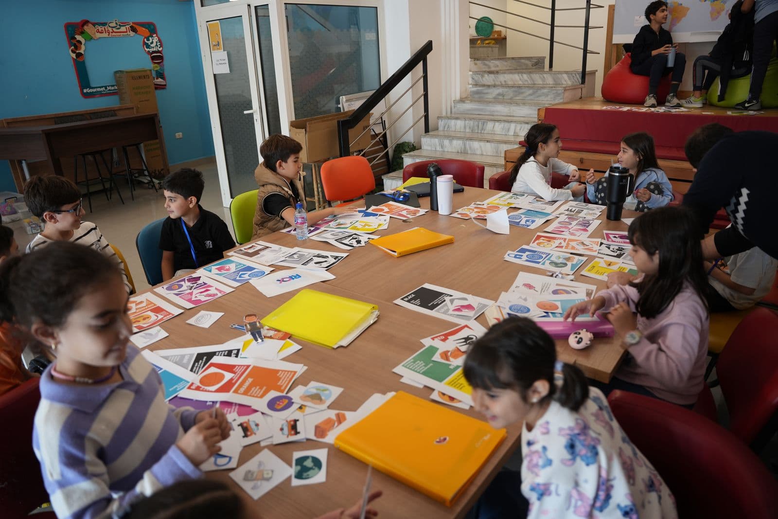 Children seated around a long table covered in colourful activity cards and folders, Youngpreneurs Academy