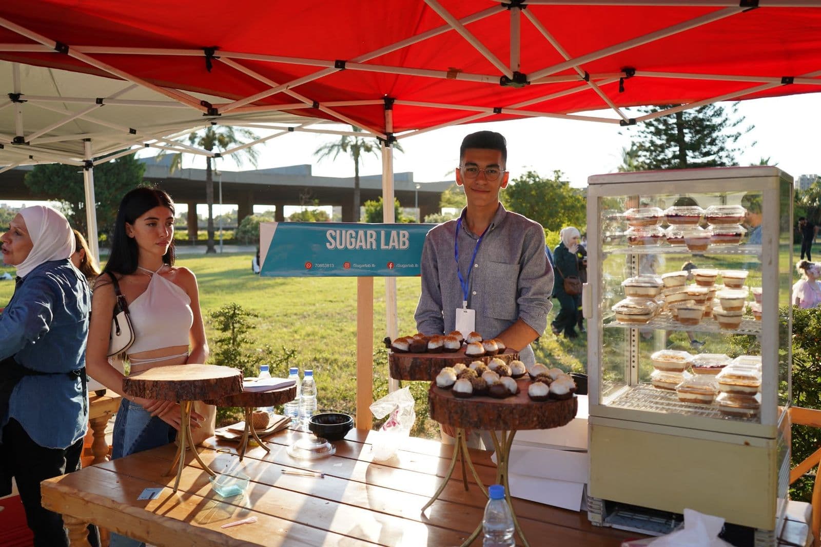 A vendor behind an outdoor dessert stall with a cake display case at the Gourmet Market