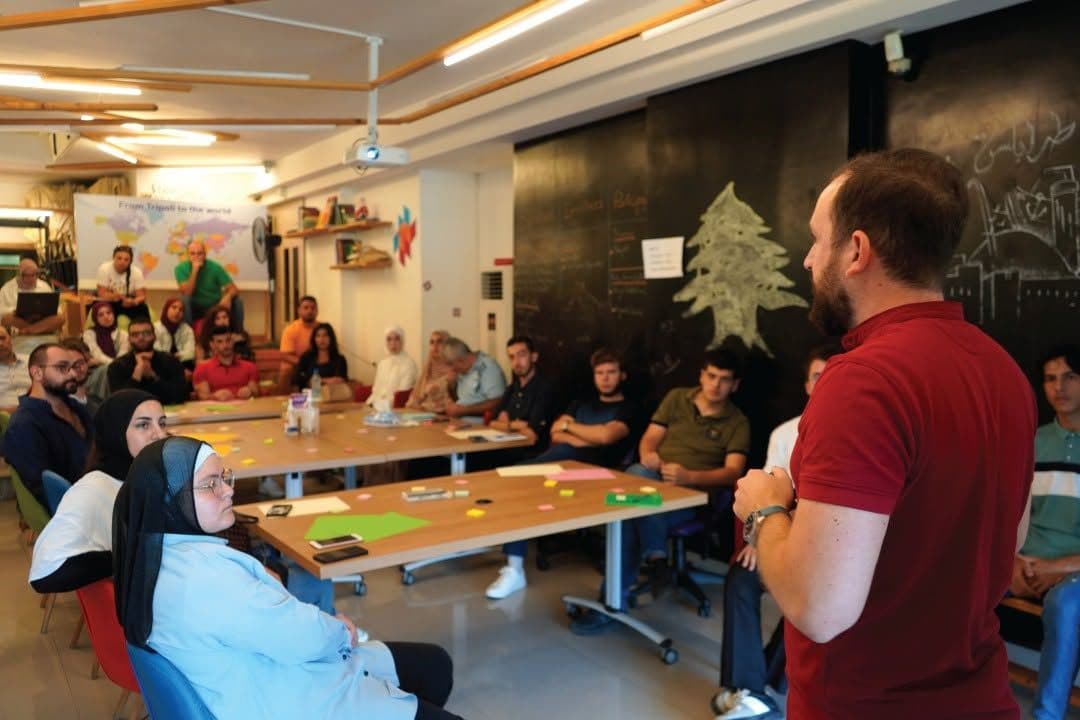 A facilitator stands and speaks to participants seated around workshop tables
