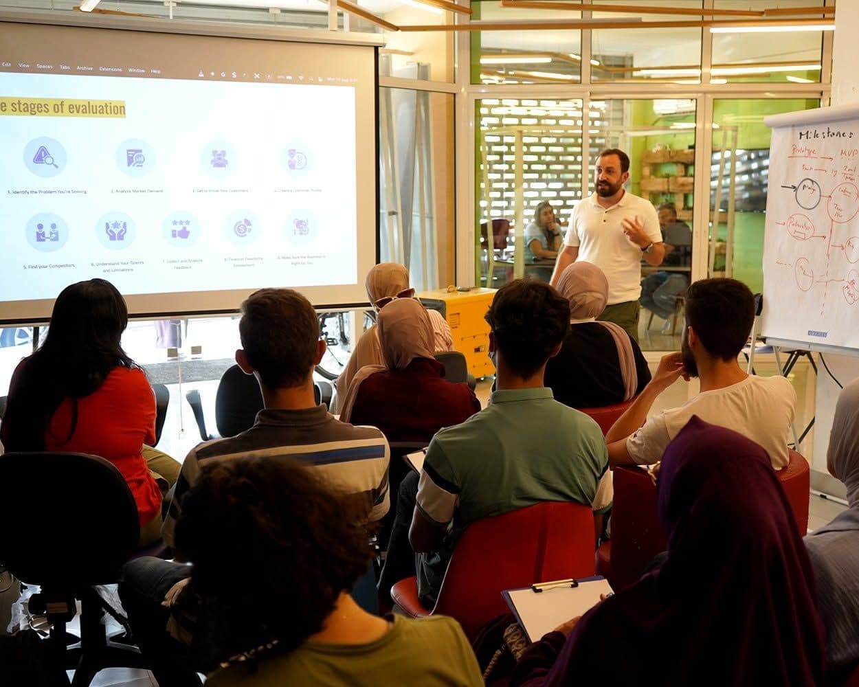 A presenter explains a projected slide to a full room of workshop participants