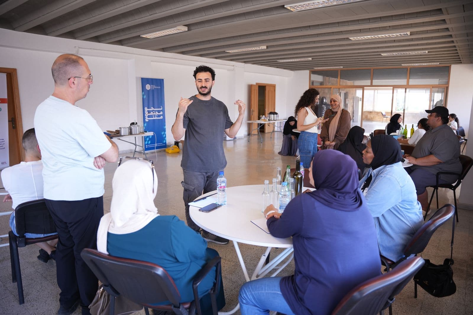 A facilitator speaks to community members seated at round tables during a Haretna workshop