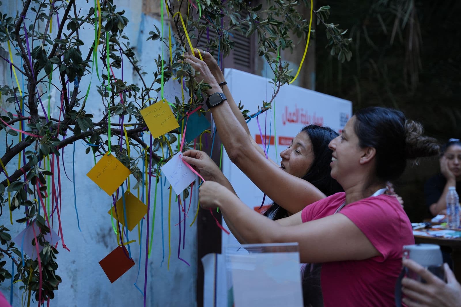 Women hang coloured paper notes on ribbons tied to a tree during a Haretna activity