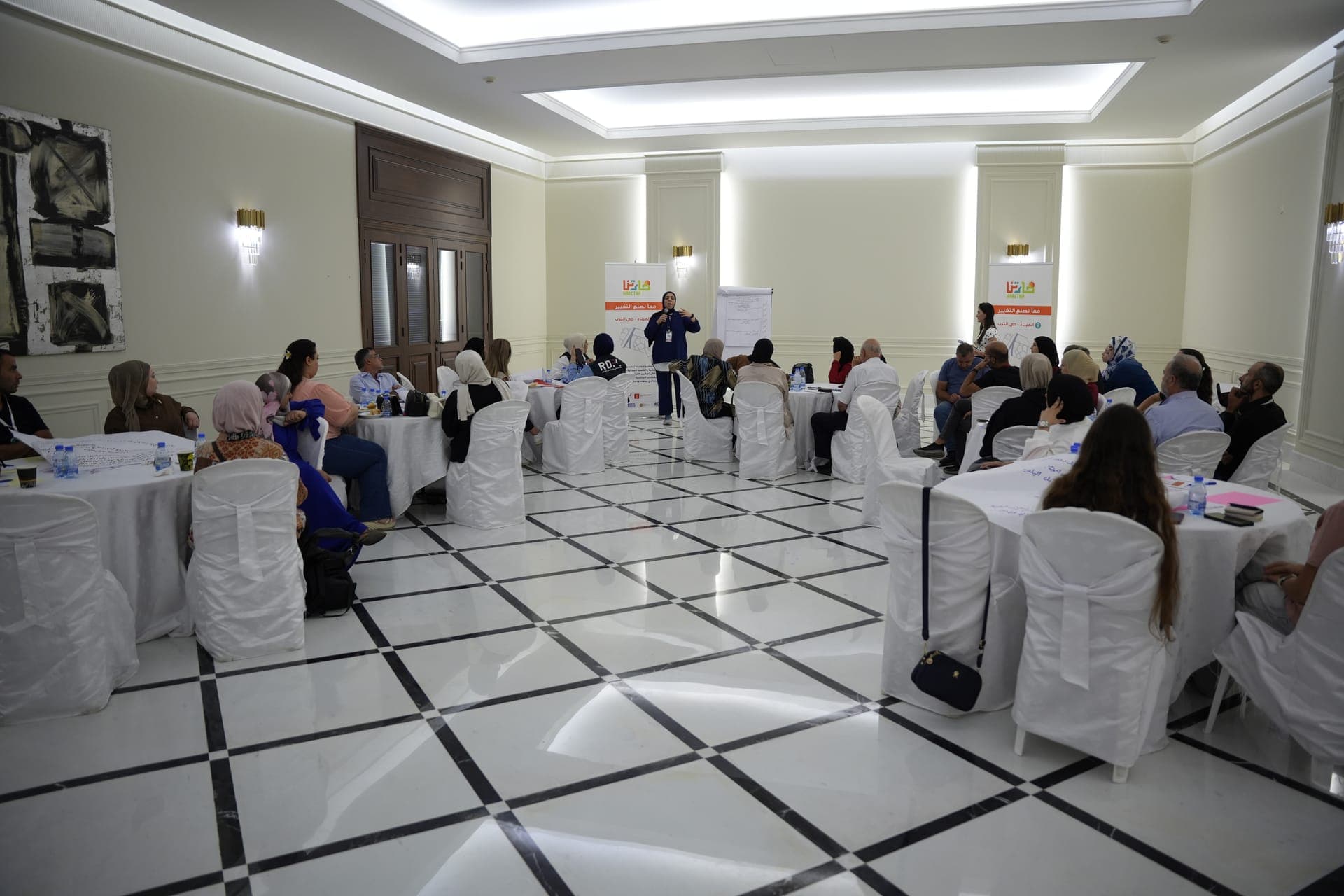 Community members seated at round tables in a large hall during a Haretna session